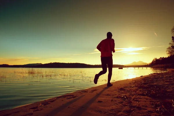 Grand homme avec coupe-vent rose et casquette sombre courir sur la plage au coucher du soleil — Photo