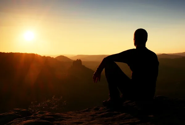 Caminante deportivo en ropa deportiva negro sentarse en la cima de la montaña y tomar un descanso. Vigilancia turística hasta el valle brumoso de la mañana . — Foto de Stock