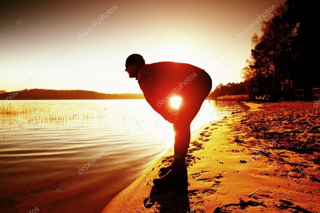 Silhouette of sport active adult man running and exercising on the beach. Calm water, island and sunset sky background.