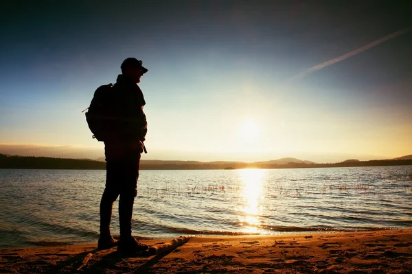 Wycieczkowicz wysoki w ciemnej odzieży sportowej z Polaków i sportowy plecak na plaży podziwiać zachód słońca na horyzoncie z niebieski niebo z chmurami. — Zdjęcie stockowe