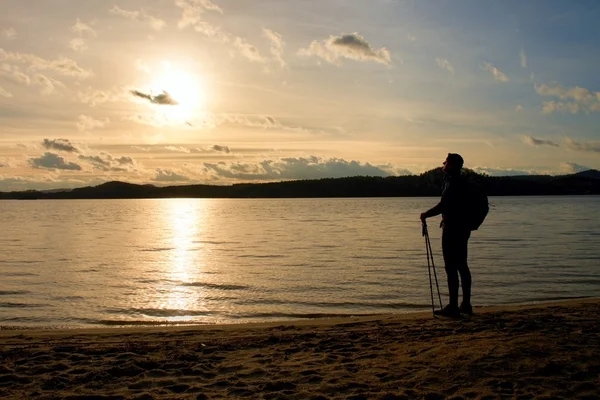 Senderista alto en ropa deportiva oscura con postes y mochila deportiva en la playa disfrutar de la puesta de sol en el horizonte. Día mágico de otoño . — Foto de Stock