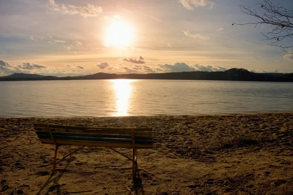 Banco vazio ao ar livre na praia do lago outono. A costa com o pôr do sol. Foto tonificada vintage com efeito de flare da lente — Fotografia de Stock