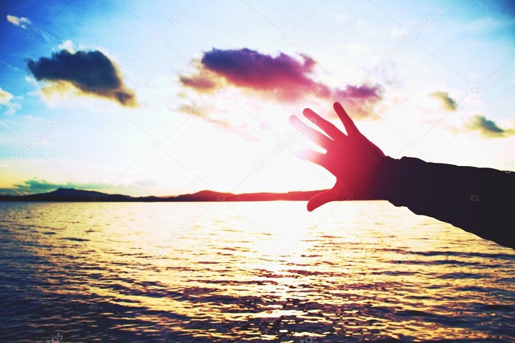 Silhouette of hand and lomg fingers try to touch the sun,  sunset above ocean . Vivid colors.