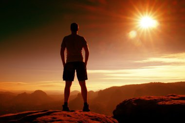 Red filter photo. Slim tourist in pants on the cliff of rock empire is watching down to valley. Hot Sun above.