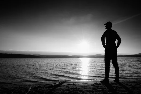 Człowiek z ręką w powietrzu. Wycieczkowicz wysoki w ciemnej odzieży sportowej z sportowy plecak stoi przy plaży podziwiać zachód słońca na horyzoncie — Zdjęcie stockowe