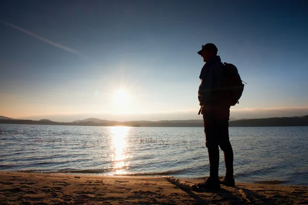 Alto escursionista in abbigliamento sportivo scuro con bastoni e zaino sportivo sulla spiaggia godersi il tramonto all'orizzonte con cielo blu con nuvole . — Foto Stock