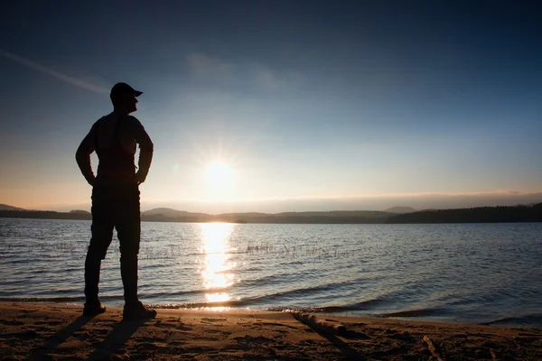 Grand randonneur en vêtements de sport sombres avec des bâtons et sac à dos sportif sur la plage profiter du coucher de soleil à l'horizon avec ciel bleu avec nuages . — Photo