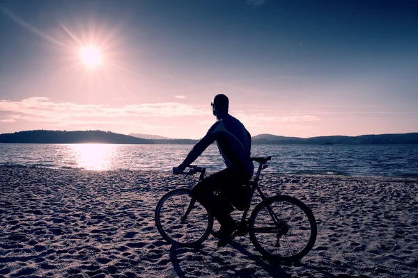 年轻男子在海滩上的日落背景和骑自行车的人轮廓在蔚蓝的天空。在湖的赛季结束. — 图库照片