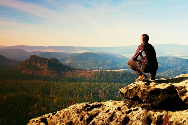 Jovem de sportswear preto está sentado na borda do penhasco e olhando para o vale nebuloso abaixo — Fotografia de Stock