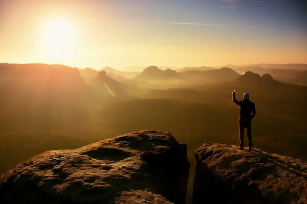 Man takes photos with smart phone on peak of rock empire. Dreamy fogy landscape, spring orange pink misty sunrise in a beautiful valley — Stock Photo, Image