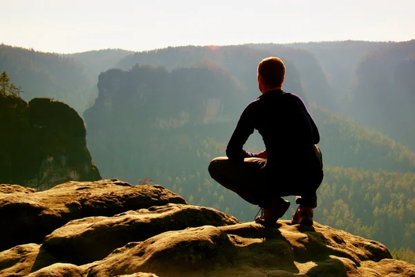 Caminante en negro en pico. Maravilloso amanecer en las montañas, niebla naranja pesada en el valle profundo. El hombre se sienta en la roca . — Foto de Stock