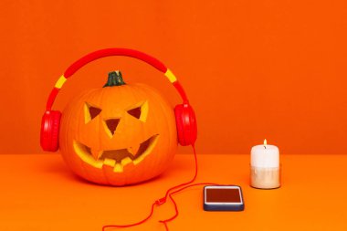 Kulaklıkla oyulmuş balkabağı telefonun yanında müzik ve turuncu masada parlak turuncu arka planda mum dinliyor.