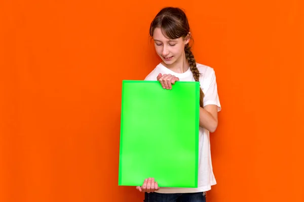 オレンジ色のスタジオの背景にバナー広告付きの明るい緑のフォルダを保持している穏やかな白人の若い女の子 広告コピースペース — ストック写真