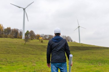 Bir erkek mühendisin arka görüntüsü, rüzgar türbini mevkiinde, sonbahar bulutlu, yeşil çayır yeşil bir plan taşıyan beyaz bir başlık takıyor.