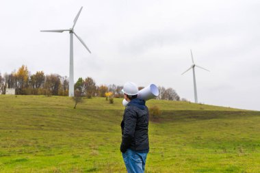 Bir erkek mühendisin arka görüntüsü, rüzgar türbini mevkiinde, sonbahar bulutlu, yeşil çayır yeşil bir plan taşıyan beyaz bir başlık takıyor.