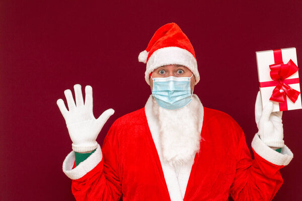 Christmas gift. Santa Claus wears medical mask holding gift box palms at tops and looking at camera while standing against red studio background