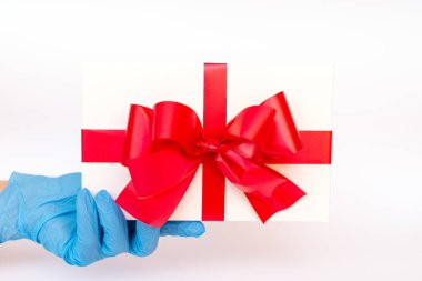 Elinde kırmızı kurdeleli bir hediye kutusu tutan meidik eldivenler vardı. Pandemik koruma. Parlak beyaz arkaplan.