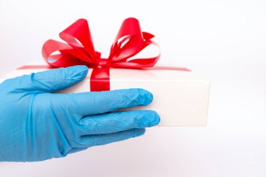 Tatil hediyesi. COVID 19 önleme. Ellerinde kırmızı kurdeleli hediye kutusu tutan meidik eldivenler var. Pandemik koruma. Parlak beyaz arkaplan.