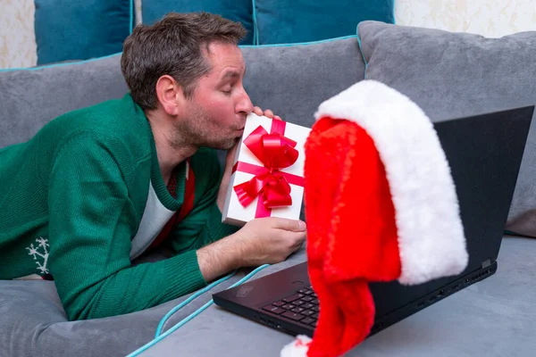 Male kisses gift. virtually exchanging Christmas gifts and having video call over a laptop at home on the couch.