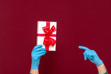 Yeni yıl 2021. Noel kutlaması, koronavirüsten korunma. Mavi eldivenli erkek elleri hediye kutusunu işaret ediyor. Kırmızı stüdyo arka planı..