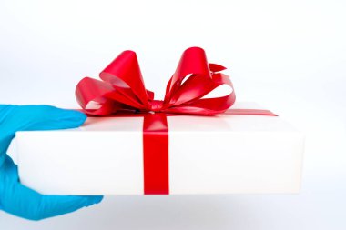 Elinde kırmızı kurdeleli bir hediye kutusu tutan meidik eldivenler vardı. Pandemik koruma. Parlak beyaz arkaplan.