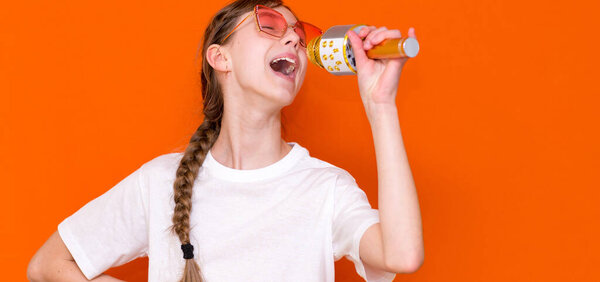 Happy young beautiful girl in sunglasses singing with microphone on orange studio background