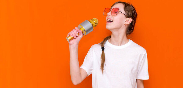 Happy young beautiful girl in sunglasses singing with microphone on orange studio background