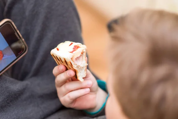 家でホットドッグを食べながらスマホで遊んでいる空腹の少年 屋内ショット クローズアップ — ストック写真