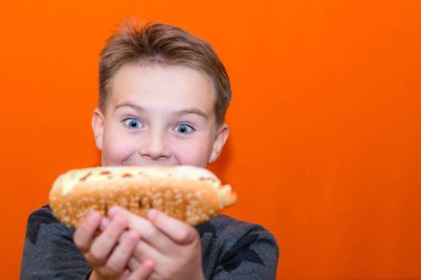Şaşırmış 10 'lu yaşların çocuğu büyük bir sosisli sandviç ısırmak istiyor. Kapalı turuncu stüdyo arka planda..