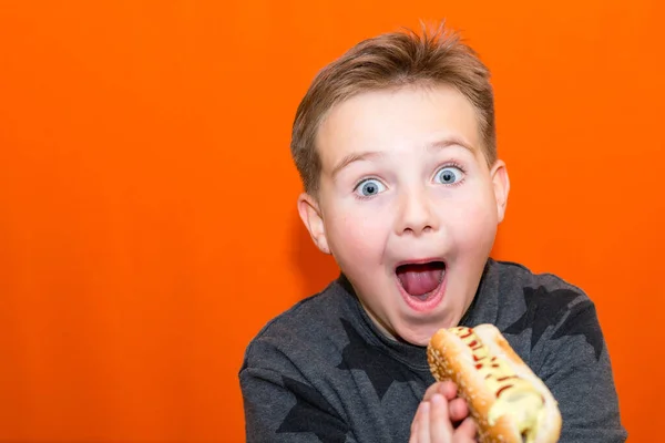 驚き10代の男の子は大きなホットドッグをかむしたいです クローズアップオレンジスタジオ屋内の背景 ボディ正 — ストック写真