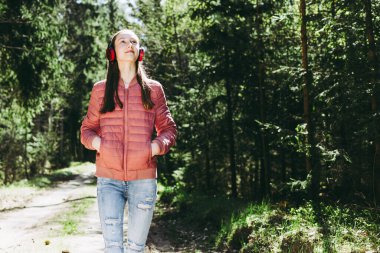 Sevimli, neşeli, sıska bir genç kız ormanda müzik dinlerken yaz, bahar günü orman ya da park manzarası seyrediyor..