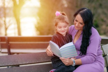 Güzel küçük kız annesiyle kitap okuyor ve gülümsüyor. Yaz, arka plandaki parkı bulanıklaştırdı. Bir kadın kızına kitap okuyor ve günbatımı parkında sıcak bir akşamın tadını çıkarıyor.