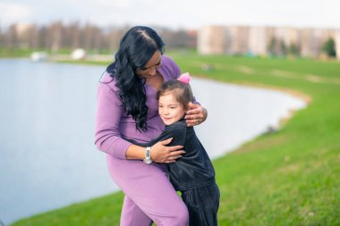İyi hafta sonları. Gülümseyen beşli kızların annesine sarılma portresi. Birlikte parkta oynuyoruz. Şık gündelik kıyafetler. Güneşli bir yaz günü. Açık hava çekimi. Şehir gölü yakınlarında.