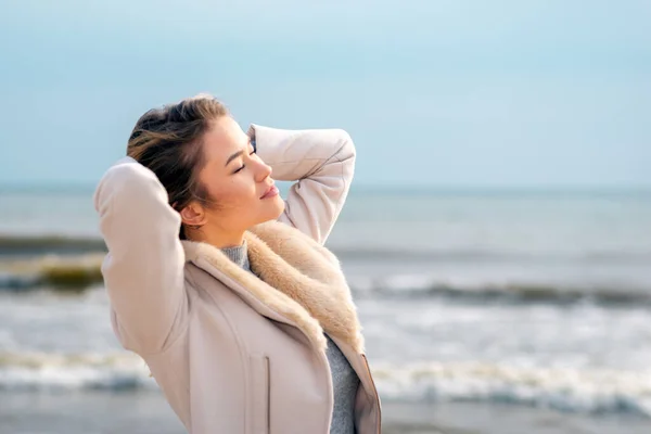 Relaxed woman, arms rised, enjoying spring sun, on a beautiful beach. Young lady feeling free, relaxed and happy. Concept of vacations, freedom, happiness, enjoyment.