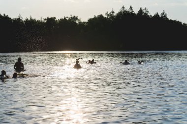 Günbatımında gölün arka planında yaşayan insanların siluetleri. Bir grup mutlu çocuk yaz gölü suyunda sahildeki iskeleden eğleniyor..