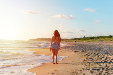 Sahilde gün batımında yalnız genç bir kadın yürüyor. Moda plajındaki kadın yaz tatilinde okyanus suyunun yanında rahatça yürüyor..