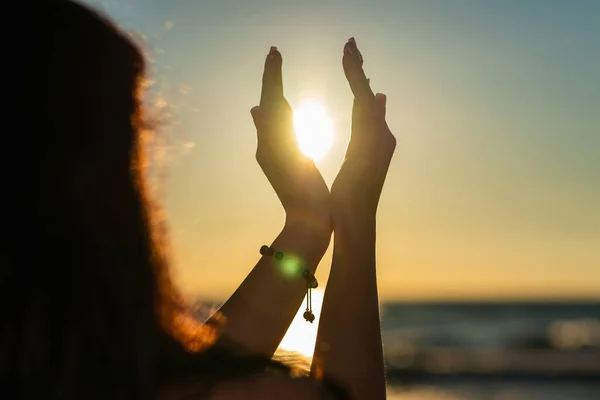 Nice summer sun solstice concept. Silhouette of young woman\'s hands relaxing, happy meditating and holding sunset against golden hour sky on the beach with natural ocean, sea background.