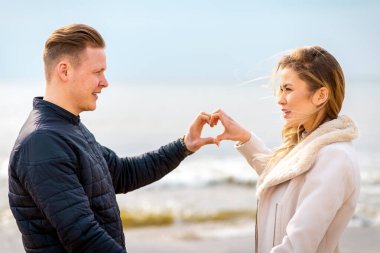 Romantik genç çift kalplerini oluşturuyor, birbirlerine bakıyor ve sahilde dururken gülümsüyorlar..