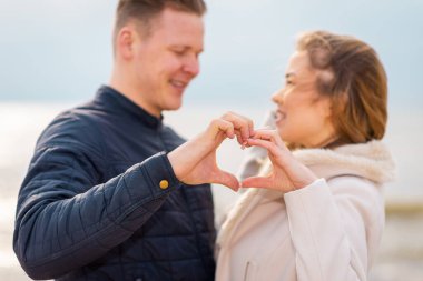 Romantik genç çift kalplerini oluşturuyor, birbirlerine bakıyor ve sahilde dururken gülümsüyorlar..