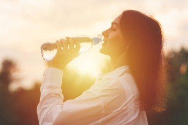 Kız plastik şişeden su içiyor. Genç beyaz kadın ormanda elinde bir şişe suyla yaz doğasında dinlenme eğitimi alırken..