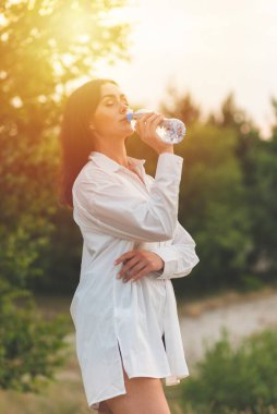 Kız plastik şişeden su içiyor. Genç beyaz kadın ormanda elinde bir şişe suyla yaz doğasında dinlenme eğitimi alırken..