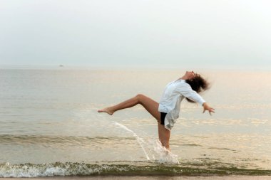 Günbatımı kumsalında su sıçratan komik genç bir kadın oynuyor. Mavi denizin kıyısında güzel, mutlu bir kadın. Eğlenceli, pozitif bir ruh hali, yaz tatili konsepti. Bulanık görüntü, hareket bulanıklığı..