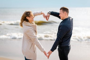 İlkbahar, sonbahar akşamları, deniz kıyısında kollarıyla kalp şeklinde kalp yapan genç çift. Elleriyle plajda kalp şekli oluşturan mutlu çift..