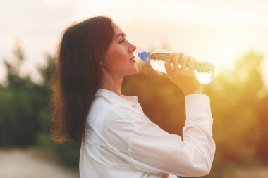 Kız plastik şişeden su içiyor. Genç beyaz kadın ormanda elinde bir şişe suyla yaz doğasında dinlenme eğitimi alırken..