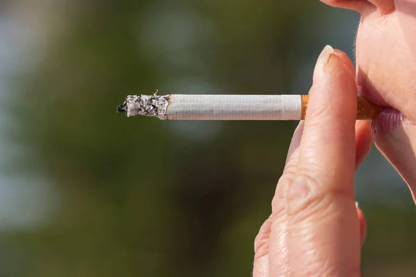 Middle age woman smoking cigarette outdoors with sun on her face. Close up of a girl with a cigarette in her mouth with space for copy or text.Blurred background.Side view portrait.