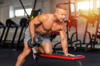 Güçlü vücut geliştirici, spor salonunda dambılla egzersiz yapıyor. Genç, yakışıklı, yetişkin bir erkek, modern spor salonunda tek kollu halterde kürek çekiyor. Boşluğu kopyala..