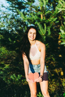 Güzel esmer, kırmızı kalp kartpostalını gösteren, rüya gibi gülümseyen, yaz güneşli bir gün. Doğada elinde kırmızı kağıt bir kalp tutan bir kadın. Yeşil arka plan..