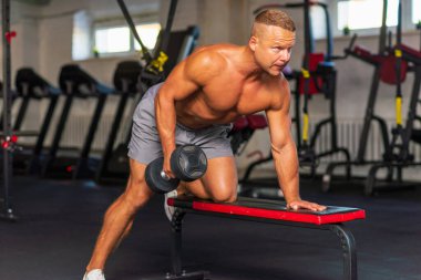Güçlü vücut geliştirici, spor salonunda dambılla egzersiz yapıyor. Genç, yakışıklı, yetişkin bir erkek, modern spor salonunda tek kollu halterde kürek çekiyor. Boşluğu kopyala..