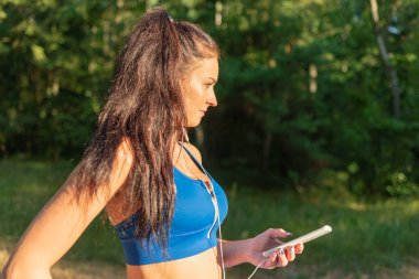 Ormanda egzersiz yapan genç ve güzel bir bayan kulaklıkla müzik dinliyor. Yaz ormanında kulaklıkla müzik dinleyen mutlu genç bir kadın. Yakından..