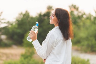 Kız plastik şişeden su içiyor. Genç beyaz kadın ormanda elinde bir şişe suyla yaz doğasında dinlenme eğitimi alırken..